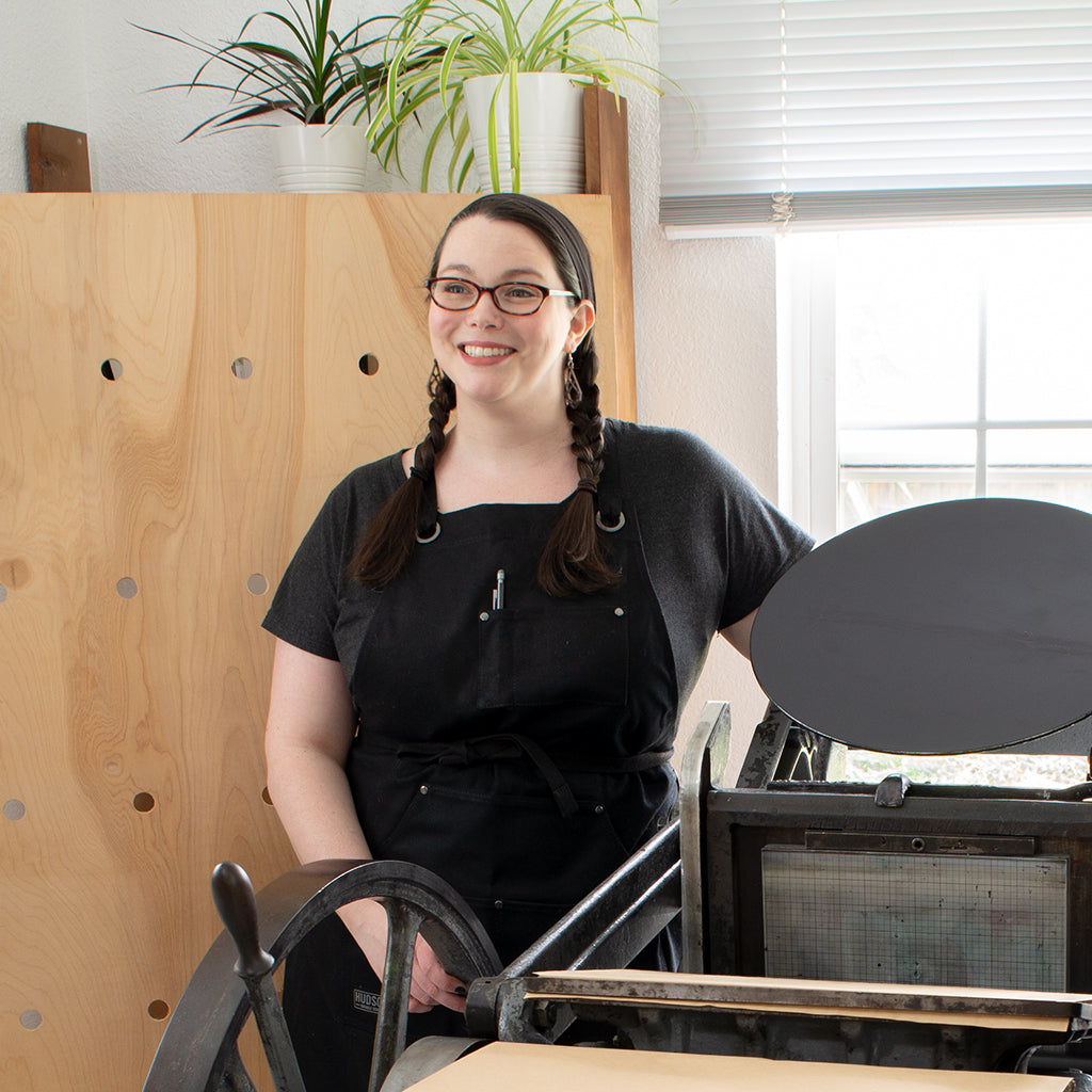 Christine, a white woman in black work apron, stands beside a printing press looking slightly up and to the left, smiling.